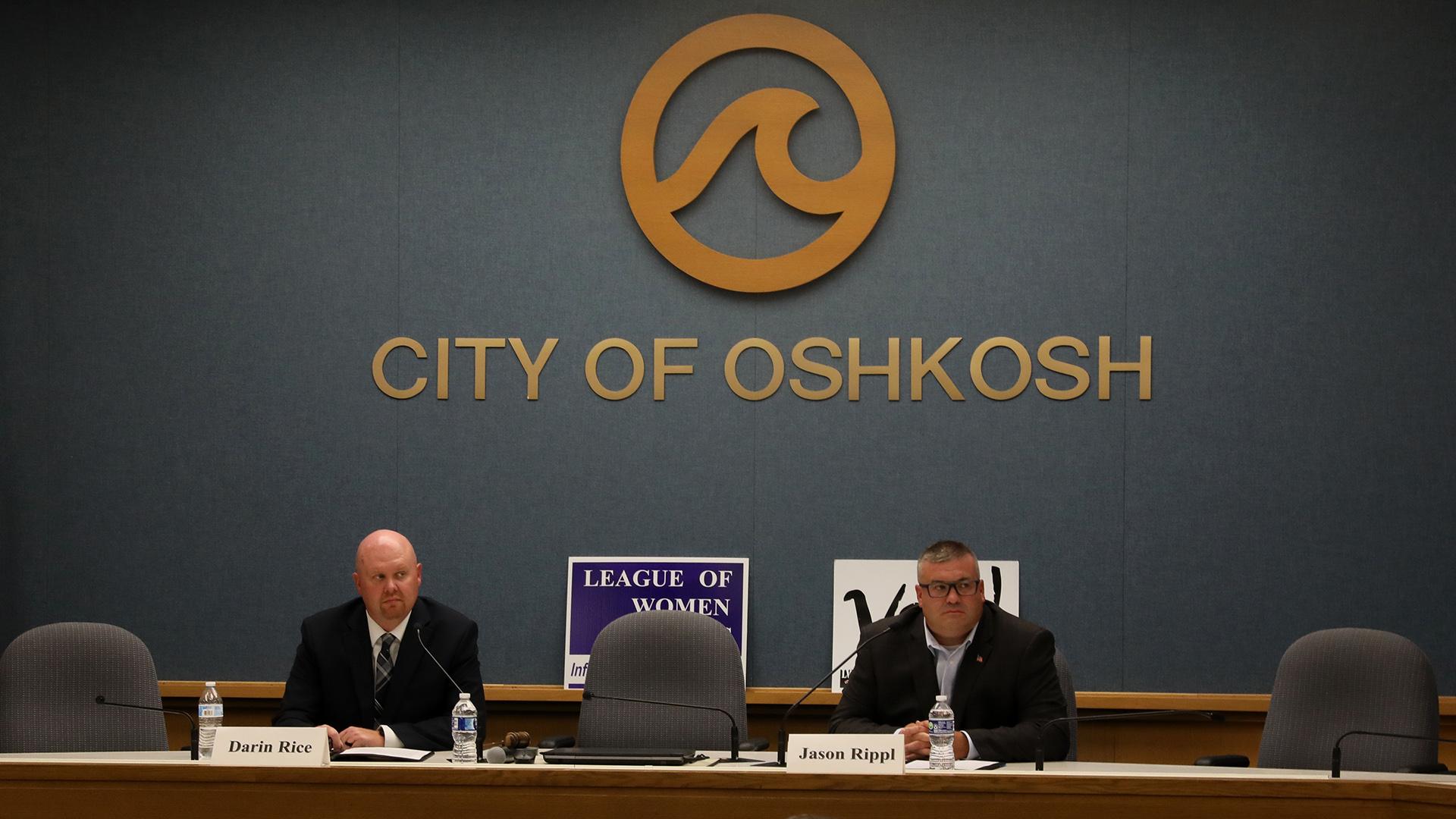 Darin Rice and Jason Rippl sit in high-backed fabric chairs behind a legislative dais while facing mounted microphones and with plastic bottles of water and paper name signs in front of them, in a room with a League of Women Voters sign displayed on a ledge in front of a wall with a logo of a water wave above a letter sign reading "City of Oshkosh."