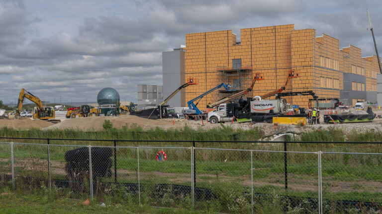 Multiple construction vehicles with extended cranes are parked in front of a multi-story building with metal siding that is under construction under a cloudy sky, with other construction vehicles and a glass geodesic dome to the side in the background, and with multiple chain-link fences in the foreground. Multiple construction vehicles with extended cranes are parked in front of a multi-story building with metal siding that is under construction under a cloudy sky, with other construction vehicles and a glass geodesic dome to the side in the background, and with multiple chain-link fences in the foreground.
