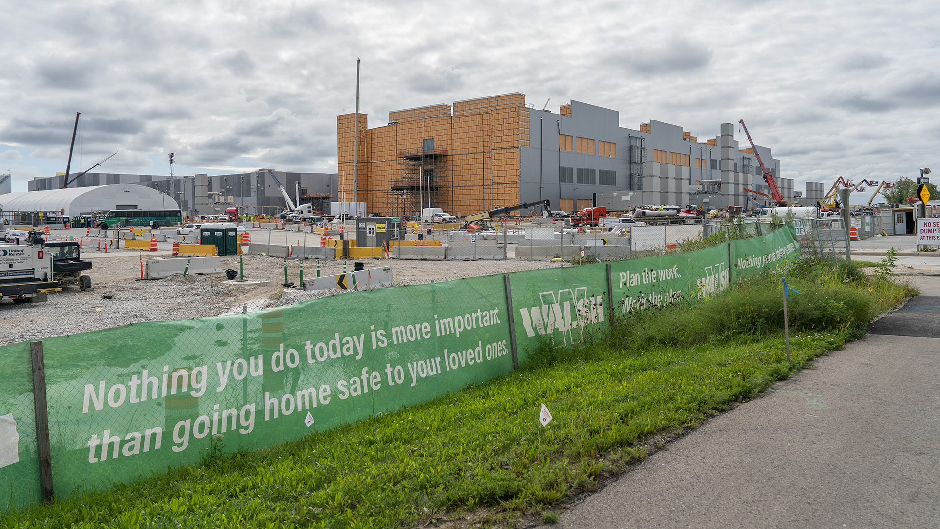 Multiple construction vehicles with extended cranes are parked in front of a multi-story building with metal siding that is under construction under a cloudy sky, with other buildings under construction in the background, and a chain-link fence covered a safety banner with the words "Nothing you do today is more important than going home safe to your loved ones" and other text in the foreground next to a paved road connected to the site.