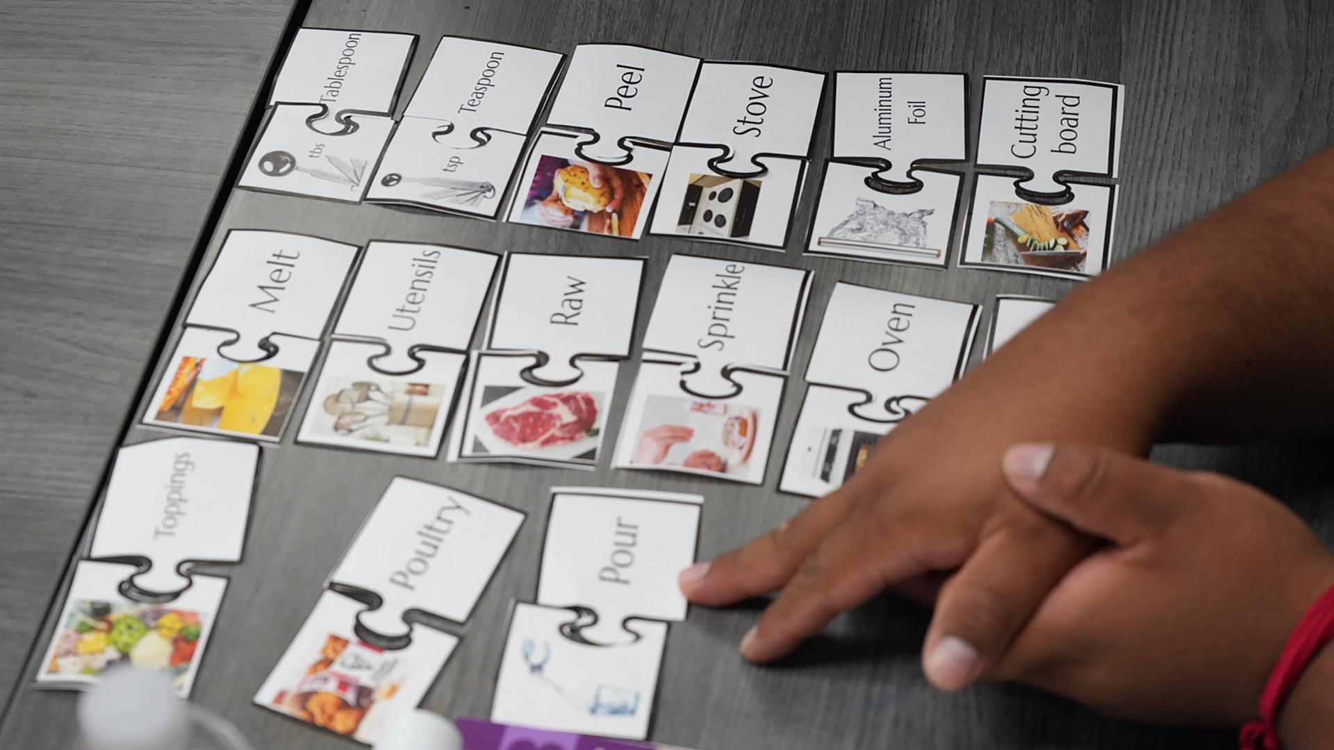 Vocabulary cards with a photo on the left matched in a puzzle piece with a word on the right are arranged in two columns on the surface of a table, with an out-of-focus pair of hands assembling a third column in the foreground.