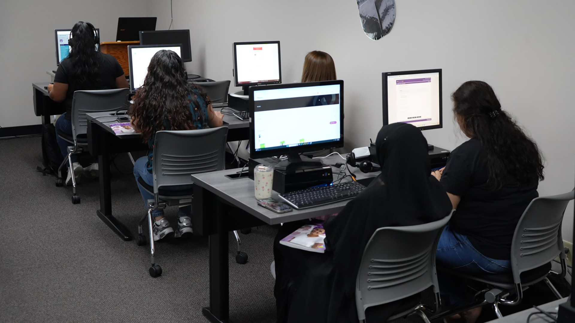 People seated in pairs on rolling chairs face side-by-side desktop computers and monitors on the surface of three desks arranged in a column next to a wall.