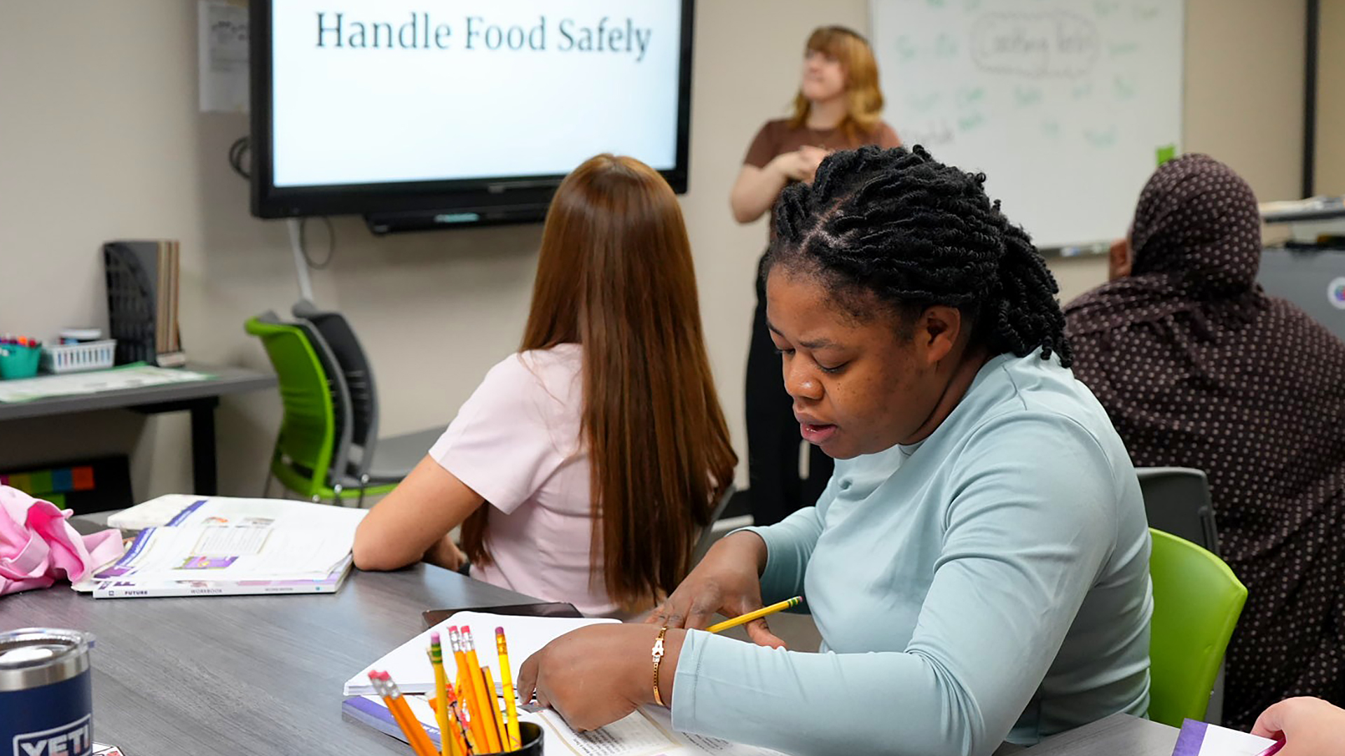 Wedcia Jean holds a pencil in her right hand while holding her right hand on an open book while seated at a table with other people seated on either side with open books and a container filled with pencils on its surface, in a room with another seated person behind her and and out-of-focus person standing in front of a video monitor with the words "Handle Food Safety" on the screen.