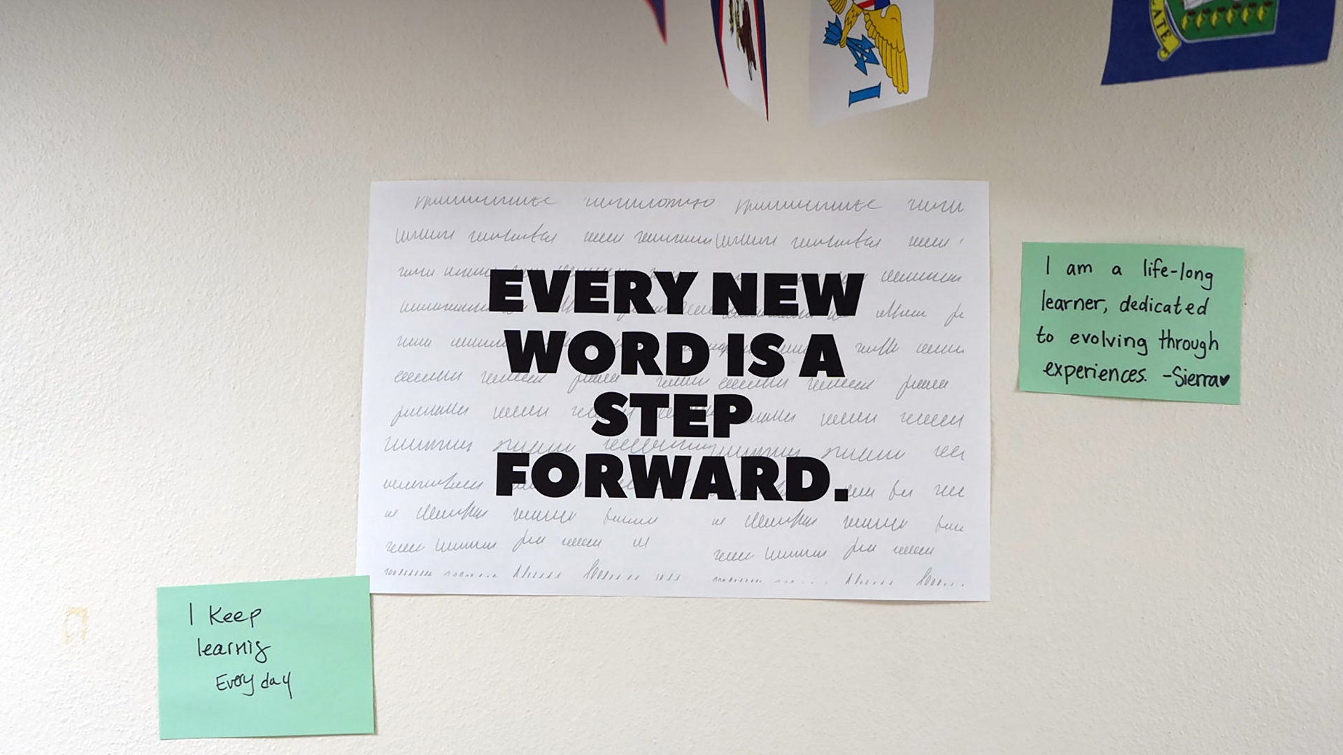 A printed paper sign with the words "Every New Word Is a Step Forward." is displayed on the surface of a wall, with two handwritten notes affixed to either side, with portions of different flags visible above.