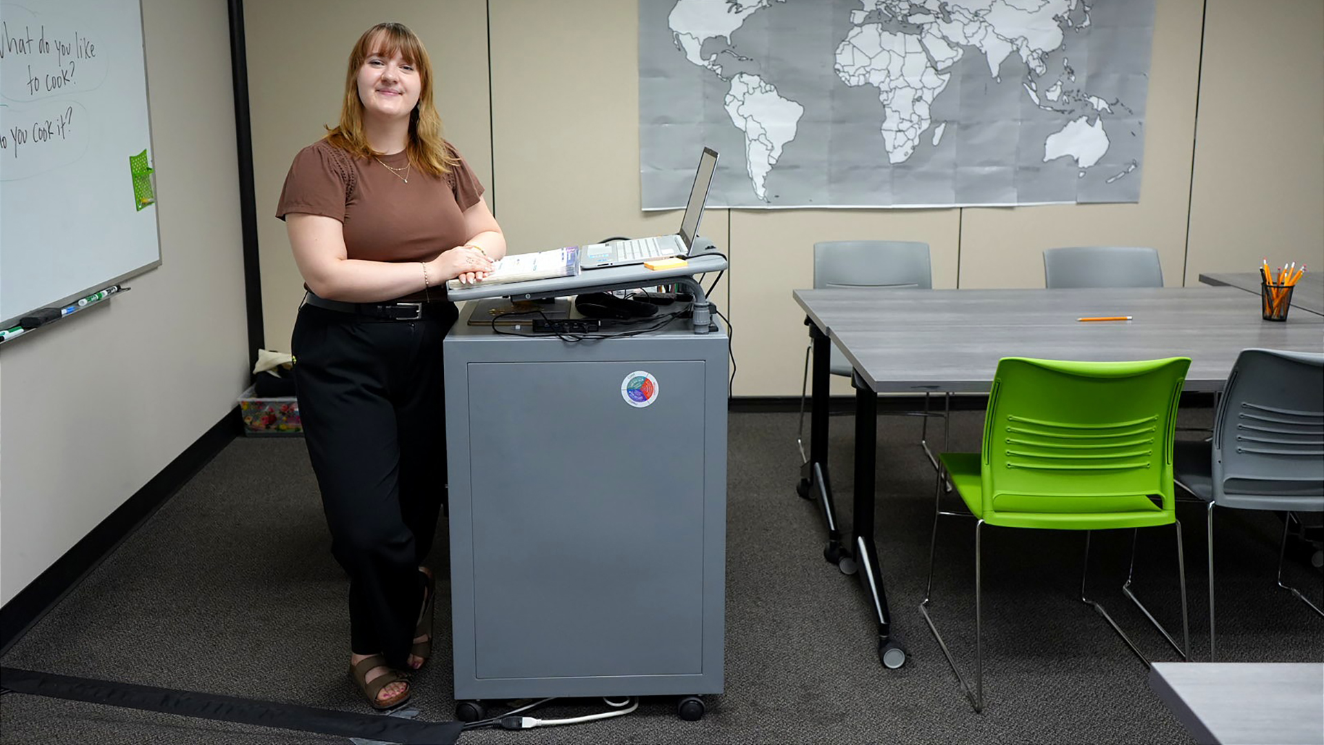 Sierra Susa poses for a portrait while standing behind a metal cabinet with a podium tray at its top with an open laptop computer on its surface, in a room with low-pile carpet, a whiteboard and world map affixed to two walls, and a rolling table with stackable chairs.