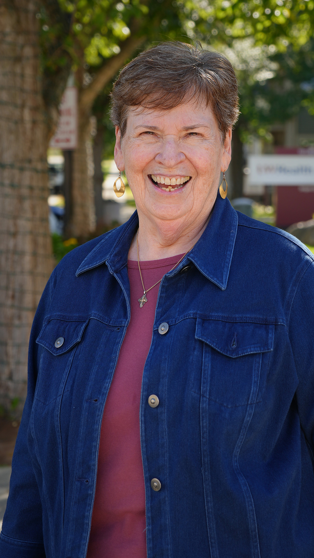 Diana Ducett poses for a portrait while standing outside under the canopy of a tree with its trunk wrapped with sting lights, with out-of-focus signs and buildings in the background.