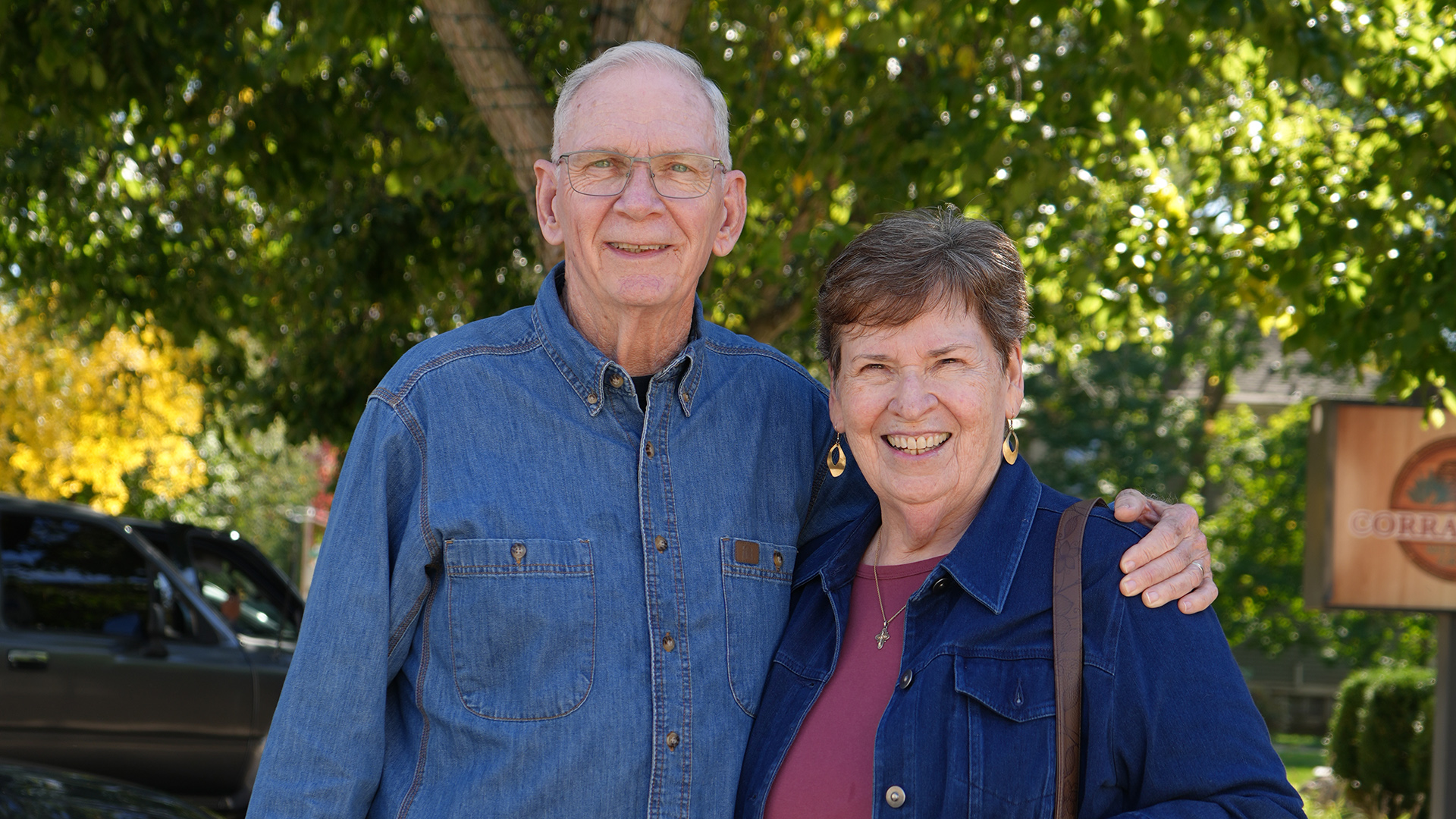 Bill and Diana Ducett pose for a portrait while standing outside under the canopy of a tree with its trunk wrapped with sting lights, with an out-of-focus business sign and parked vehicles in the background.