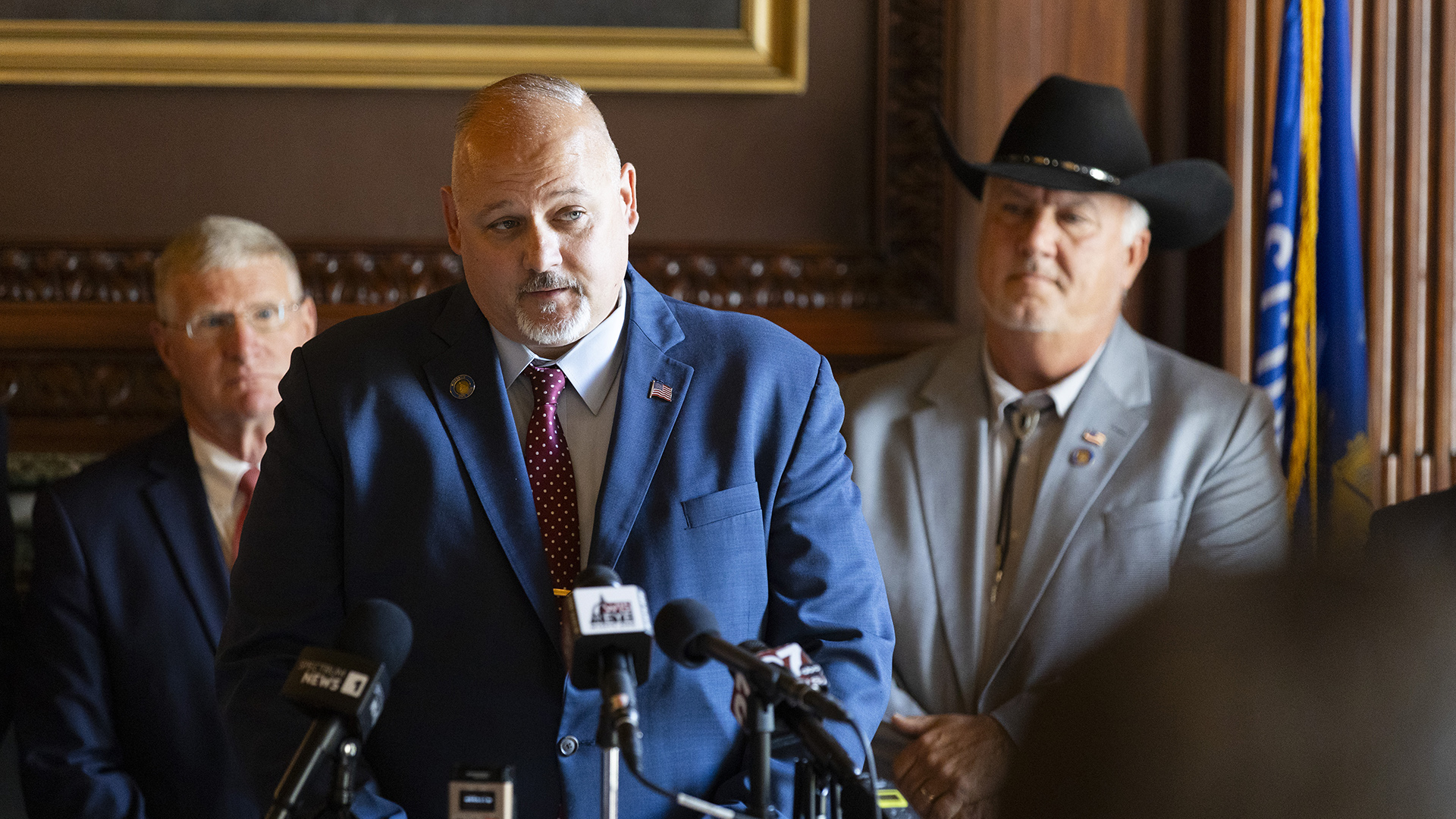 Jesse James speaks while standing behind multiple microphones with the flags of different media organizations with two out-of-focus people standing behind him in a room with a wood wall panels, the bottom edge of a framed painting and a Wisconsin flag, with the out-of-focus head of another person to the side in the foreground.