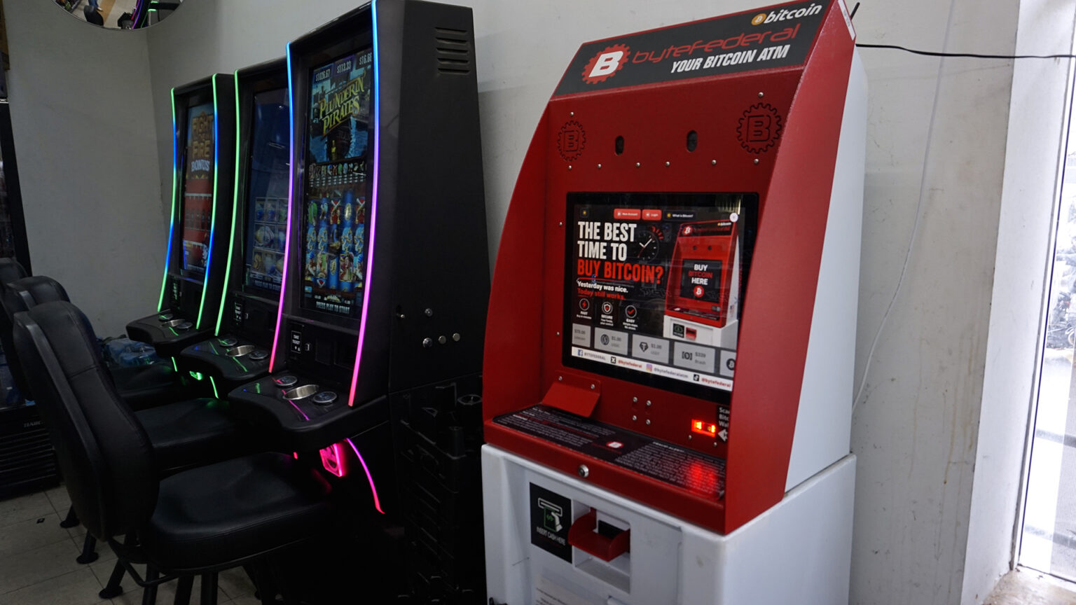 A cryptocurrency ATM kiosk with an illuminated touchscreen with the words The Best Time to Buy Bitcoin? stands next two three video gambling machines with stackable chairs facing each, in a room with a window on one side and a ceiling-mounted round mirror in a corner.
