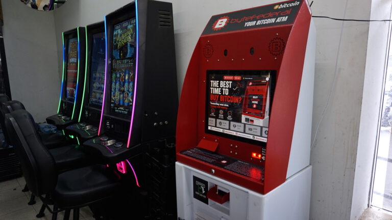 A cryptocurrency ATM kiosk with an illuminated touchscreen with the words The Best Time to Buy Bitcoin? stands next two three video gambling machines with stackable chairs facing each, in a room with a window on one side and a ceiling-mounted round mirror in a corner. A cryptocurrency ATM kiosk with an illuminated touchscreen with the words The Best Time to Buy Bitcoin? stands next two three video gambling machines with stackable chairs facing each, in a room with a window on one side and a ceiling-mounted round mirror in a corner.