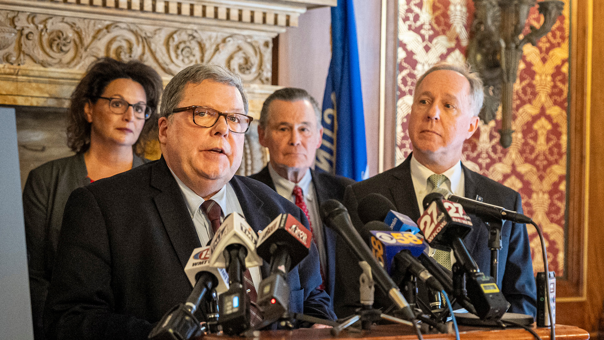 Patrick Snyder speaks while standing behind a wood podium with multiple microphones with the flags of different media organizations affixed to its surface, with Robin Vos standing to his side and two other people standing in the background, in a room with a carved fireplace lintel, the Wisconsin flag, a metal light fixture and toile wallpaper.