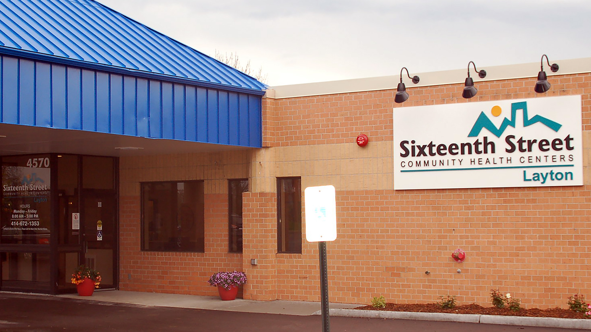 A sign with a logo of a skyline and the words "Sixteenth Street Community Health Centers" and "Layton" is mounted to the size of a brick-sided building under three spotlights mounted to the edge of the roof, with a pyramidal metal-sided awning covering a glass-walled entrance.