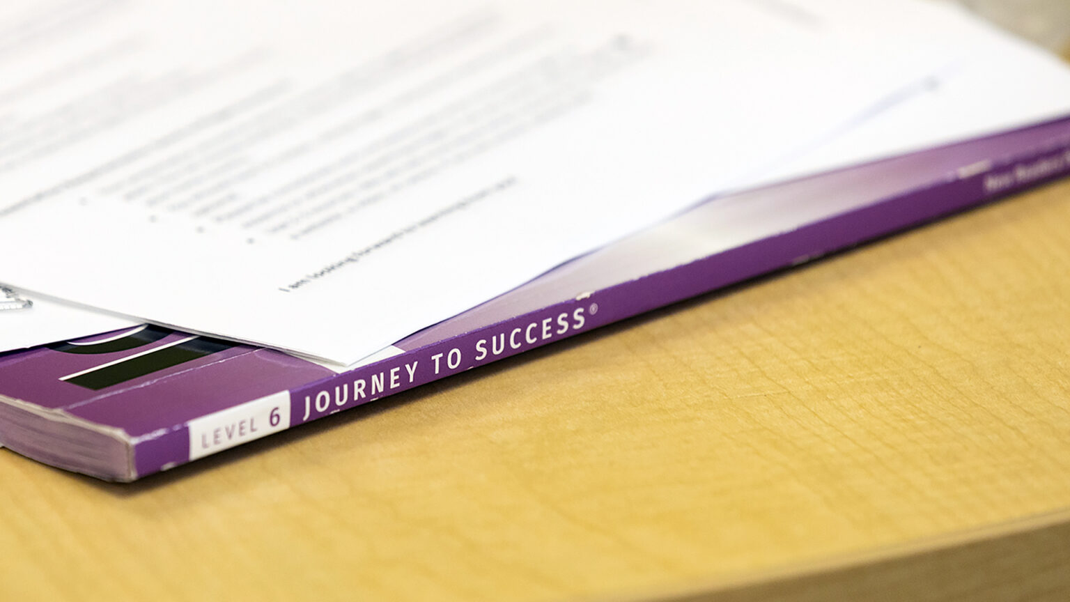 A paperbound text book with the words Level 6 and Journey to Success on its spine sits on the wood-laminate surface of a table, with multiple out-of-focus papers on top of it.