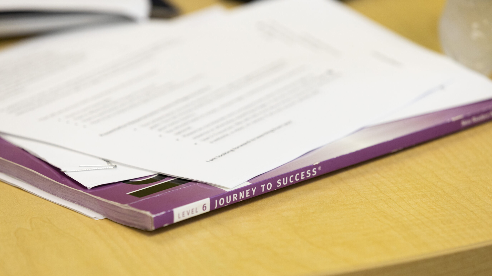 A paperbound text book with the words "Level 6" and "Journey to Success" on its spine sits on the wood-laminate surface of a table, with multiple out-of-focus papers on top of it.